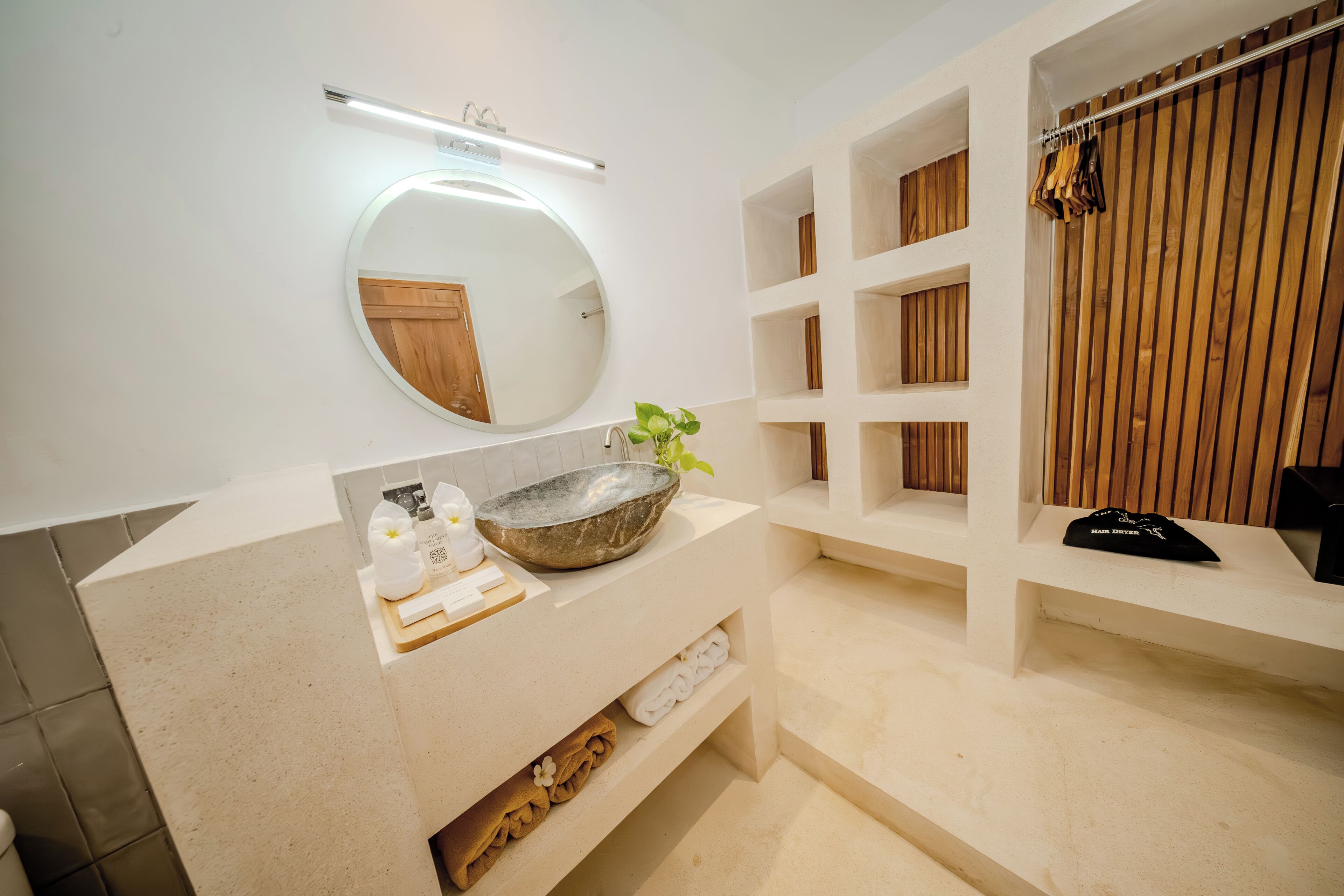 Un baño moderno con un espejo redondo, un lavabo de piedra, estantes de madera y un perchero para toallas.