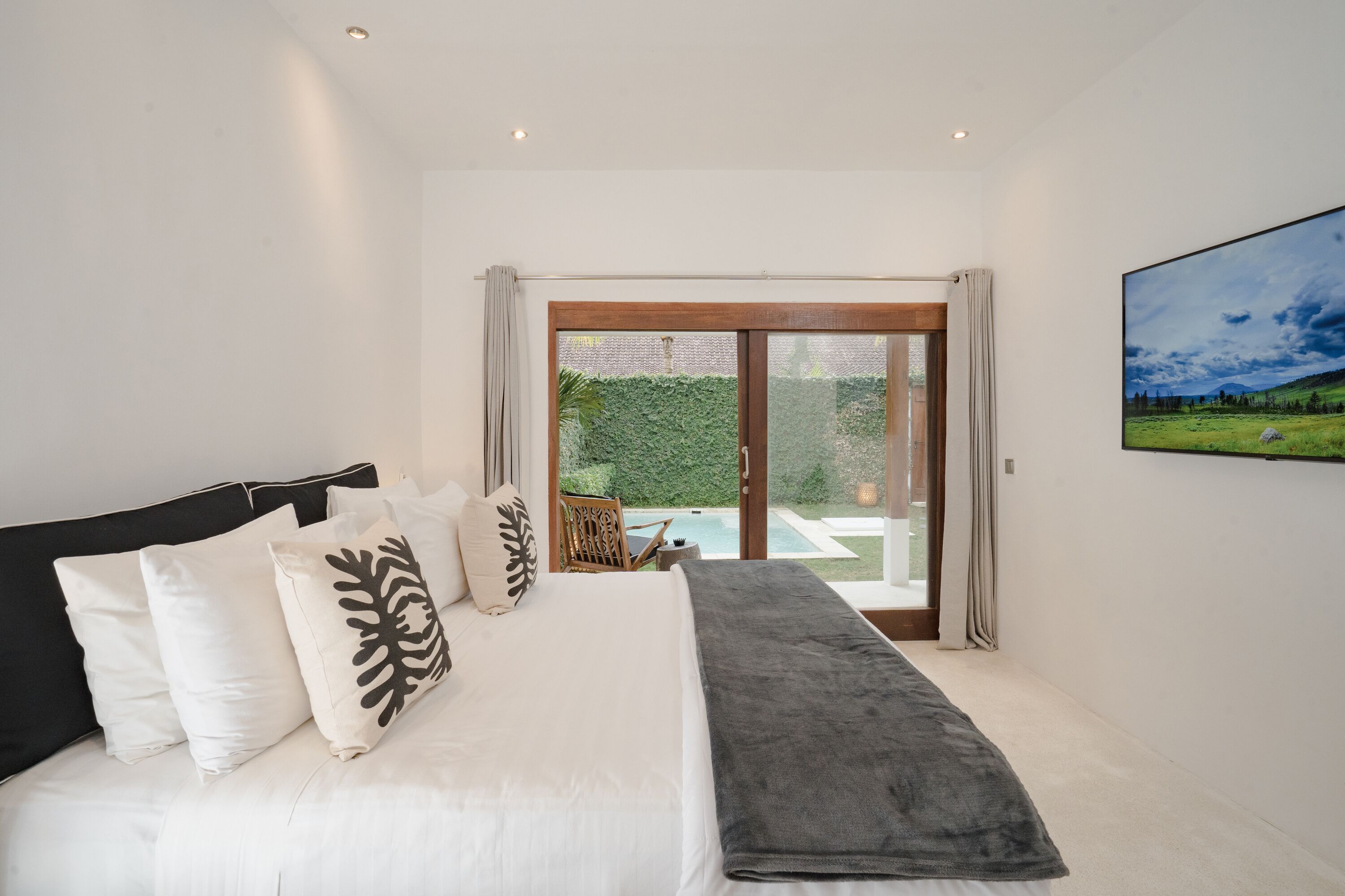A bedroom with a large bed, white bedding, a grey throw, and a view of a garden and pool through a sliding glass door.