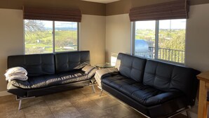 Ein modernes Wohnzimmer mit zwei schwarzen Ledersofas, einem Glastisch und großen Fenstern mit Blick auf eine grüne Landschaft.