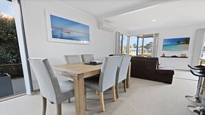 Un comedor moderno con una mesa de madera y sillas grises tapizadas, un sofá oscuro y un amplio ventanal con vistas al exterior.
