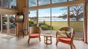 Amplia habitación con ventanas grandes que dan a un lago, con sillones de mimbre y una pequeña mesa.