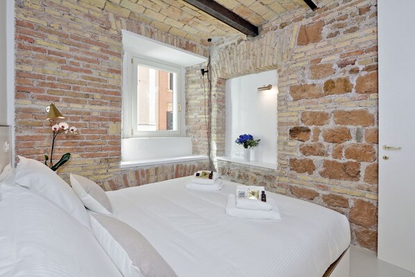 A bedroom with a brick wall, a large window, a bedside table with toiletries, and a bed with white linens.
