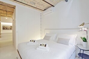 Ein modernes Hotelzimmer mit einem großen Bett, weißer Bettwäsche, einem kleinen runden Tisch mit einer Lampe und einem Blumentopf.