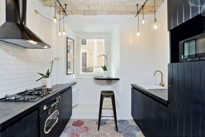 A modern kitchen with black cabinets, a gas stove, and a black range hood. There is a small stool and a window with a view of the outside.