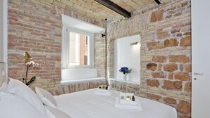 Ein Schlafzimmer mit Backsteinwand, großem Fenster, Nachttisch mit Pflegeutensilien und einem Bett mit weißen Bettwäsche.