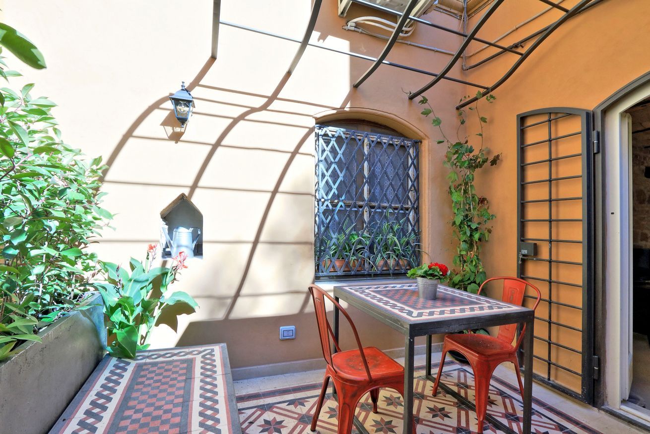 Un piccolo patio esterno con tavolo e sedie, circondato da piante e un muro decorativo.
