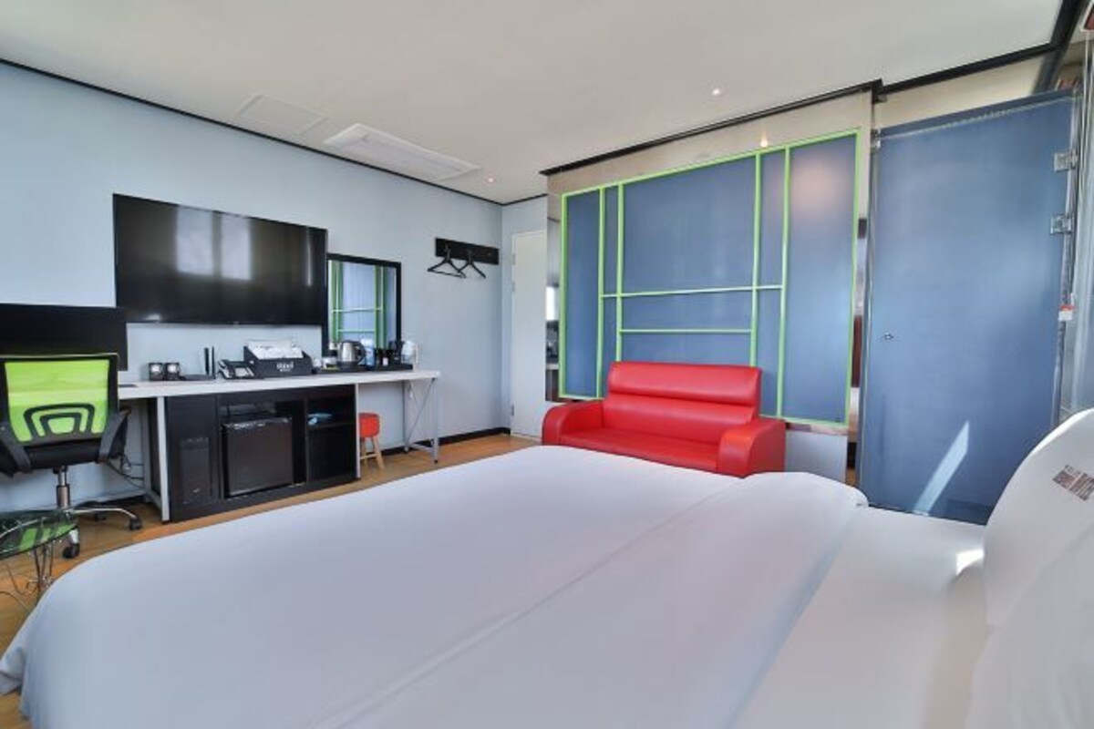 Quarto de hotel moderno com uma cama grande, uma escrivaninha com computador, uma cadeira vermelha e uma janela ampla com moldura azul.
