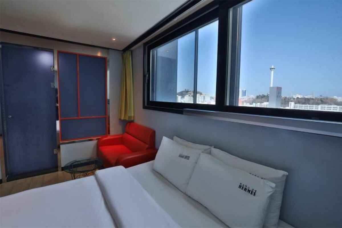 Une chambre d’hôtel avec un lit, une chaise rouge, une fenêtre donnant sur la ville et une porte avec un cadre rouge.
