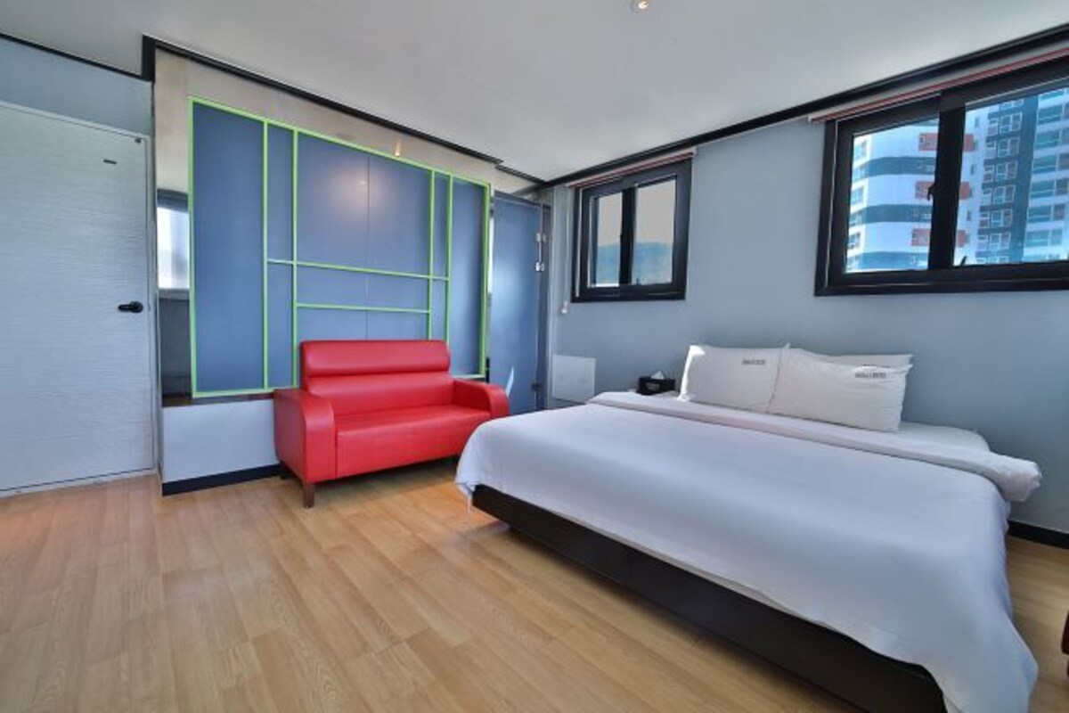 Une chambre d’hôtel moderne avec un grand lit, un fauteuil rouge et une vue sur la ville par la fenêtre.