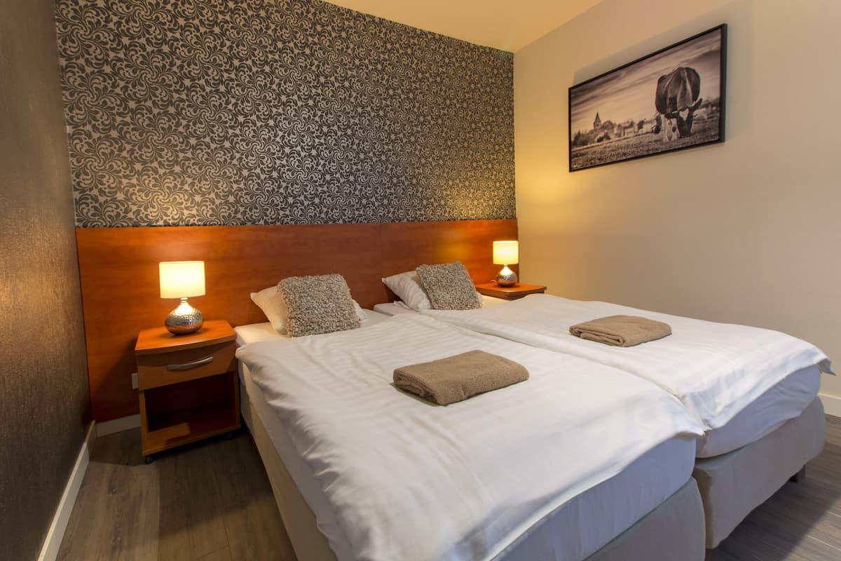 Un lit double avec du linge de lit blanc et deux oreillers, une table de chevet en bois avec une lampe, et un papier peint à motifs sur le mur.