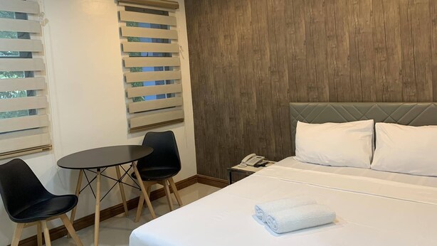 A hotel room with a bed, two chairs, a small table, a phone, and a window with blinds. - Maine City (Manila)