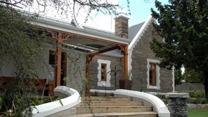 A stone house with a covered porch, steps leading to the entrance, and a stone wall to the right. - 1A St Aidans Guest House (Makhanda)