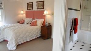 A bedroom with a bed, bedside lamps, a nightstand, and a bathroom with a shower and tiled floor. - 1A St Aidans Guest House (Makhanda)