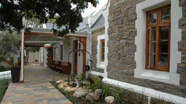 A stone building with a covered walkway, wooden benches, and a stone wall. - 1A St Aidans Guest House (Makhanda)