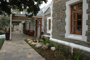 A stone building with a covered walkway, wooden benches, and a stone wall. - 1A St Aidans Guest House (Makhanda)