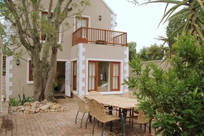 A two-story building with a balcony, a dining area with a table and chairs, and a garden with a tree and plants.