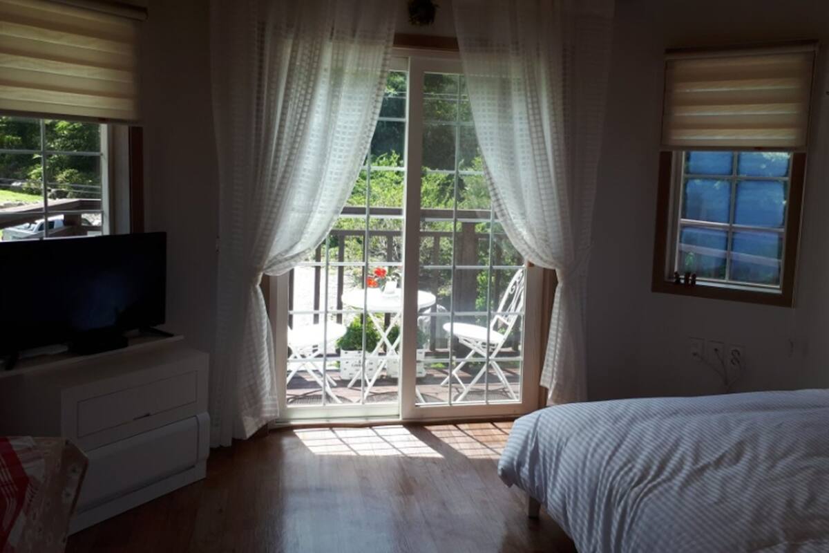 A bedroom with a bed, a TV on a dresser, a sliding glass door to a balcony, and a window with blinds.