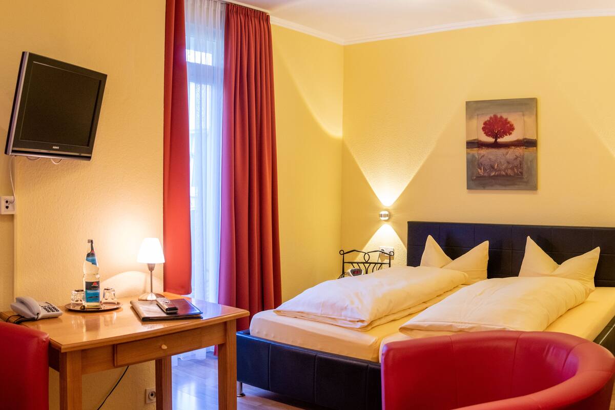 A hotel room with a bed, a desk with a telephone and a bottle, a red chair, a TV mounted on the wall, and a painting.