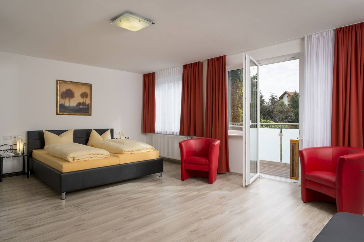 A modern bedroom with a bed, two red chairs, a painting, and a view through the glass door.