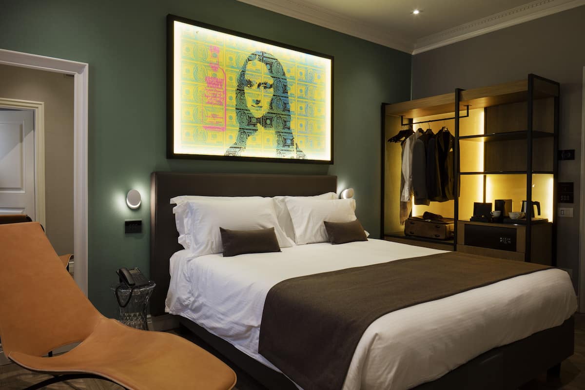 A modern hotel room with a large bed, a seating area, a wardrobe, and a wall-mounted artwork.