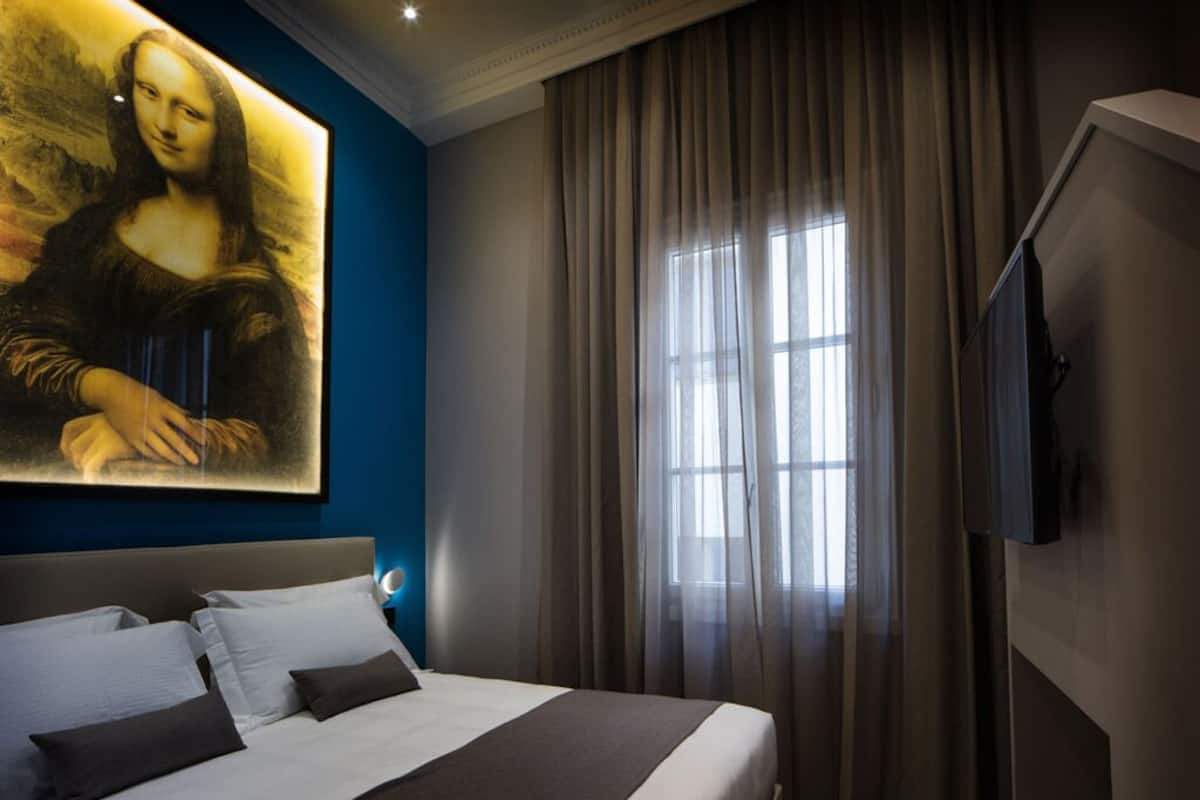 A hotel room with a large painting of the Mona Lisa on the wall, a bed with white and dark bedding, a large window with sheer curtains, and a flat-screen TV mounted on the wall.