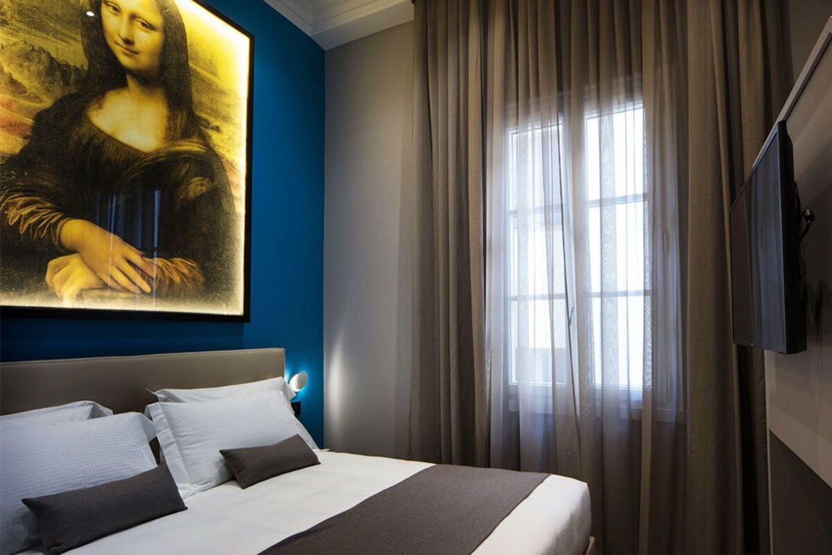 A hotel room with a large painting of the Mona Lisa on the wall, a bed with white and dark bedding, a large window with sheer curtains, and a flat-screen TV mounted on the wall.