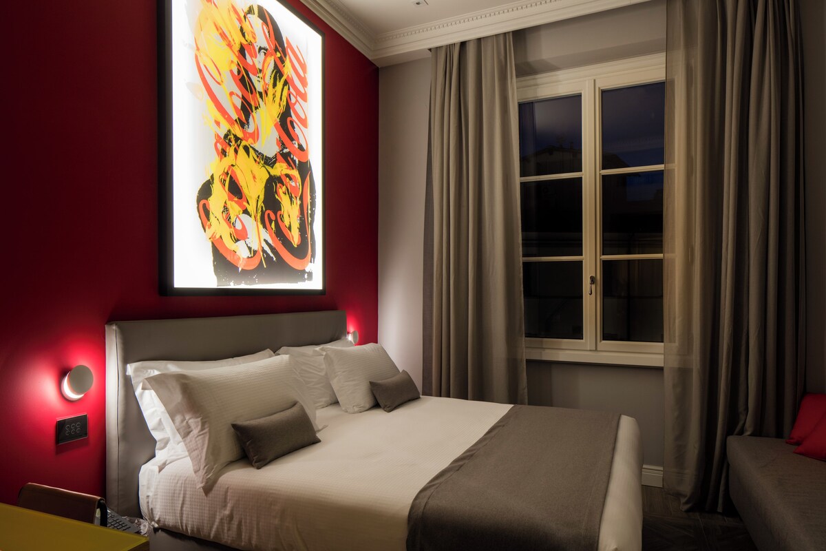 A hotel room with a large bed, a window with curtains, and a wall-mounted artwork.