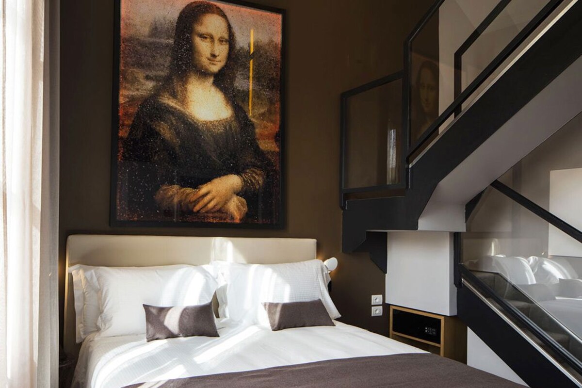 A modern hotel room with a large painting of the Mona Lisa on the wall, a bed with white linens, and a staircase leading to an upper level.
