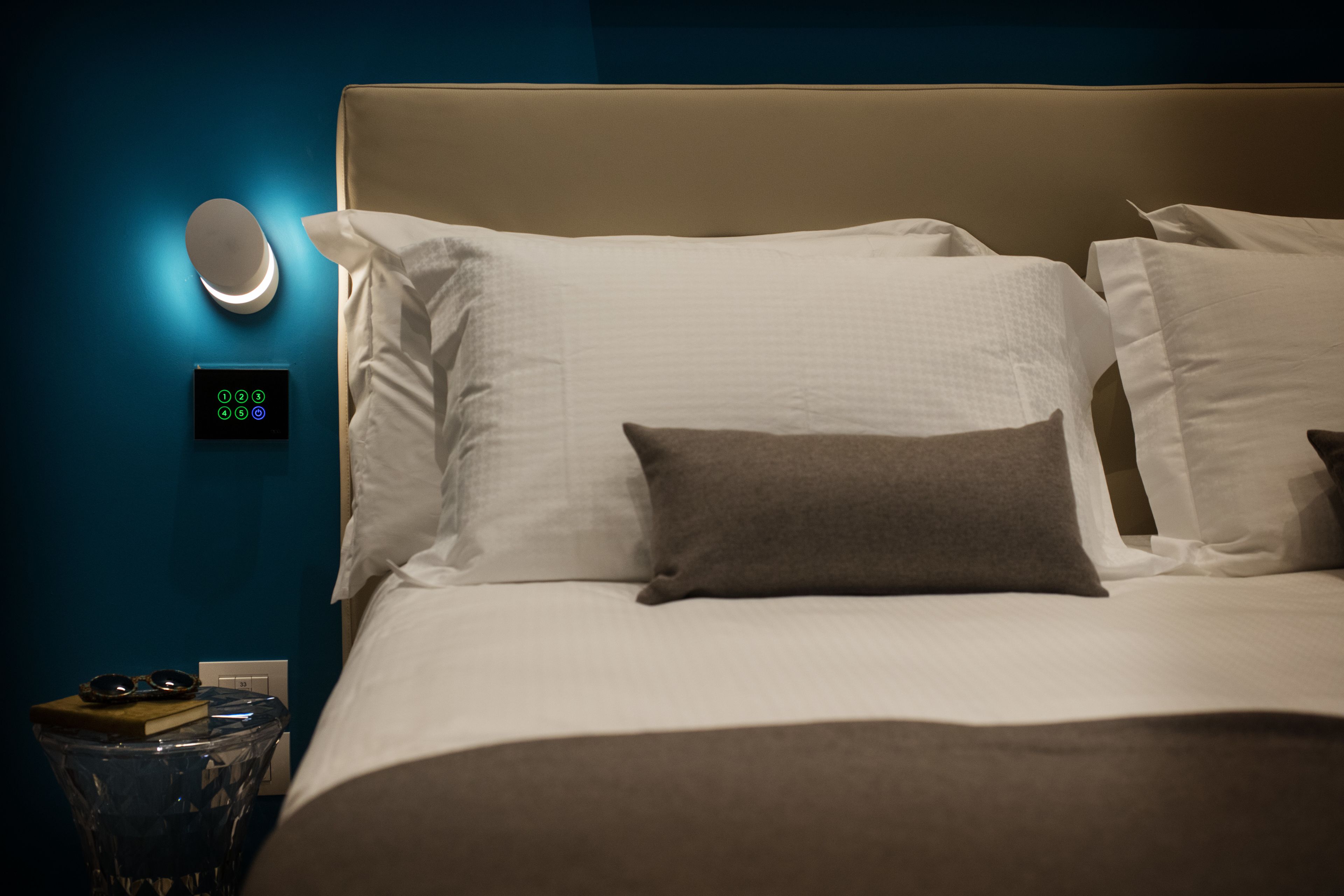 A hotel room with a bed, pillows, a bedside table with a book and glasses, a wall-mounted light, and a control panel.