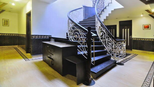 A grand staircase with ornate metal railings and a black handrail, leading to an upper floor. - Hotel Maya Cottage (New Delhi)
