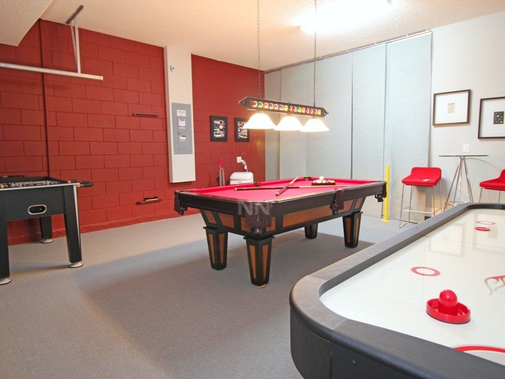 Ein Billardtisch mit roter Spielbahn, ein Kicker- und ein Shuffleboard-Tisch in einem Raum mit roten Backsteinwänden und einer weißen Decke.