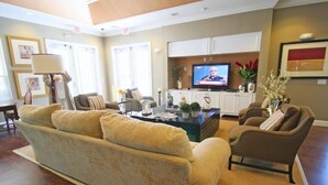 Un salon spacieux avec un grand canapé beige, une table basse et un téléviseur. On y trouve deux fauteuils et une petite table ornée d’objets décoratifs. Le sol est recouvert de parquet, et de grandes fenêtres sont équipées de stores.