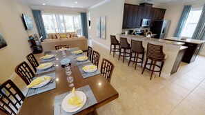 A dining area with a long table set for six, a kitchen with dark cabinets, and a living area with a sofa and TV. - Aco231263 - The Encore Club Resort - 6 Bed 6.5 Baths Villa (Kissimmee)
