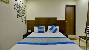 A neatly made bed with a blue and white bedspread, two pillows with blue and white stripes, a wooden headboard, a nightstand with a phone, and a door with a brown frame. - D Inn (New Delhi)