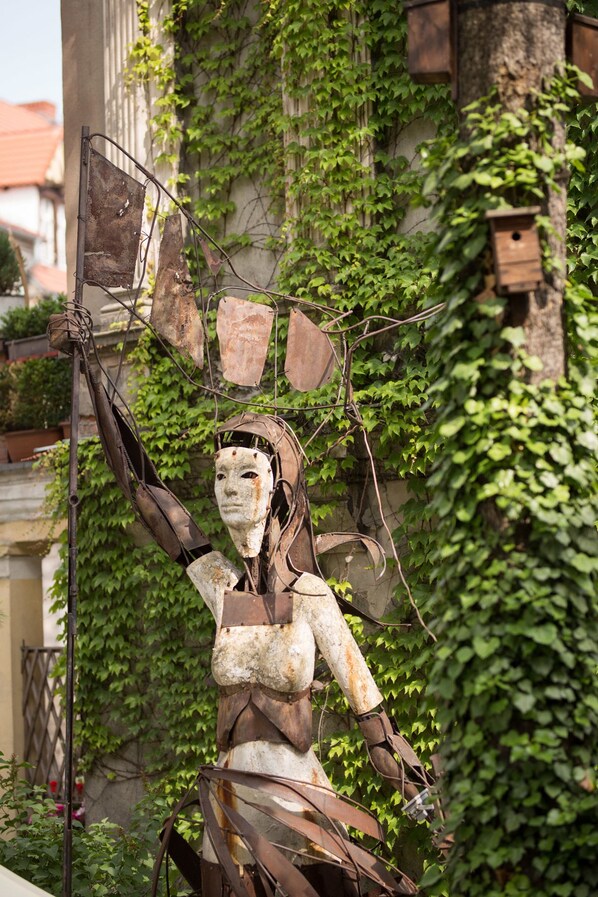 Eine Skulptur einer Figur mit erhobener Arm, umgeben von Grünpflanzen und einem Gebäude im Hintergrund.