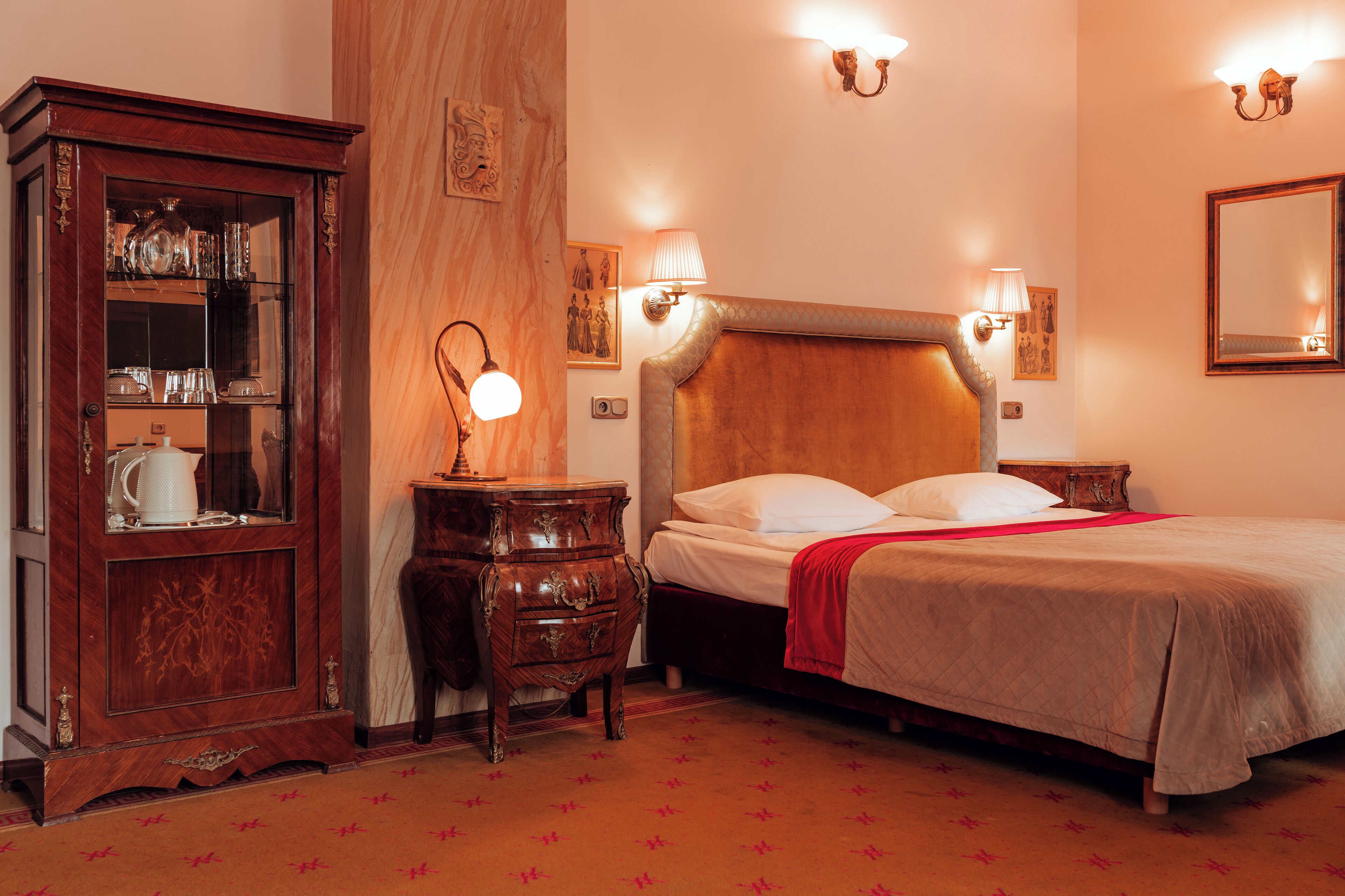 Une chambre d’hôtel avec une tête de lit en bois, un lit recouvert de draps blancs, un plaid rouge, une armoire en bois dotée de portes en verre et des lampes fixées au mur.