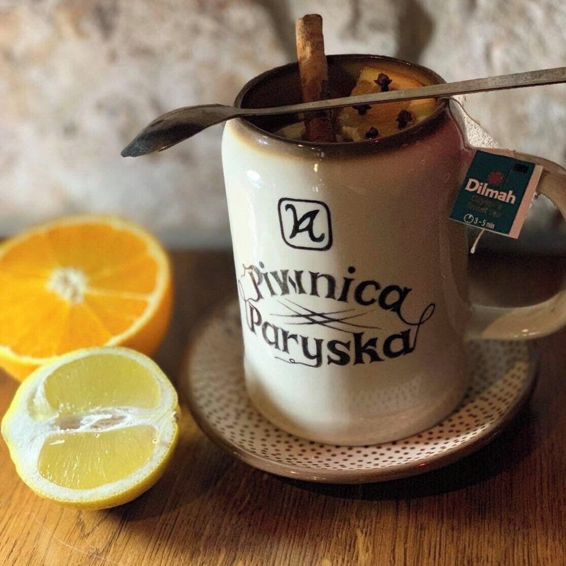 A mug with a spout and a spoon, labeled "Piwnic Paryska," containing a beverage and a cinnamon stick.