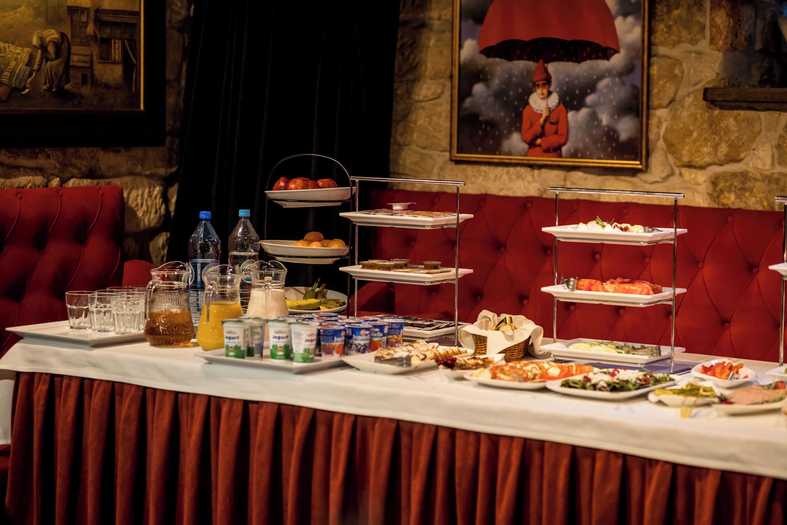 A buffet table with various food items and beverages, set against a red upholstered wall with a framed painting.