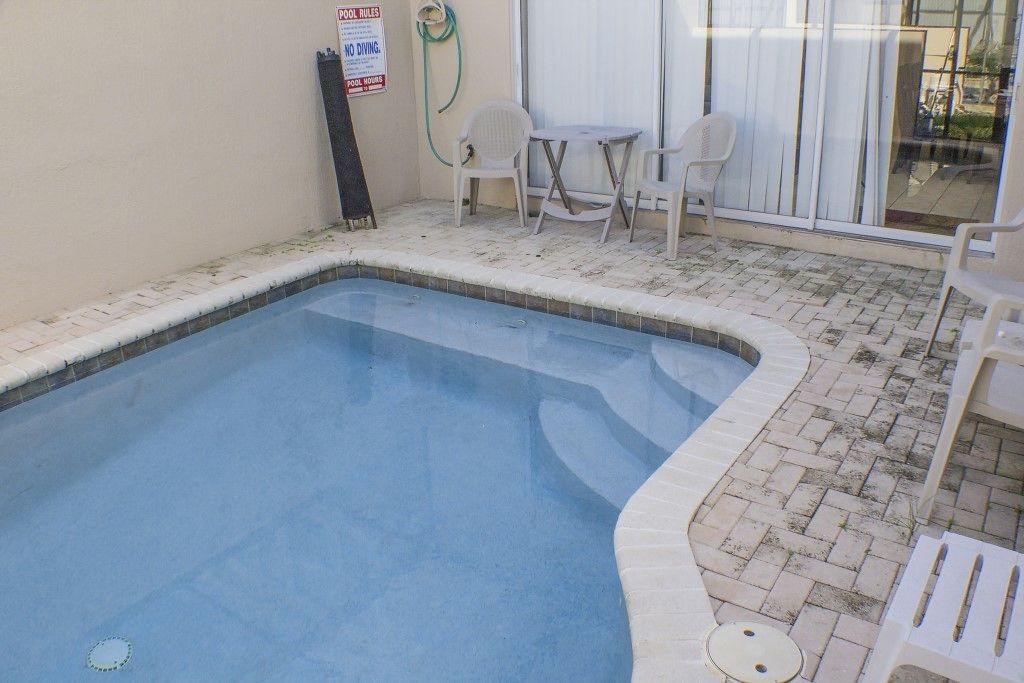 En liten inomhuspool med en kakelbeklädd kant, en skylt med poolregler och en slang. Det står två vita plaststolar och ett bord nära en skjutdörr av glas.