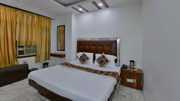 A hotel room with a large bed, a bedside table, a nightstand, a mirror, a window with curtains, and a ceiling with recessed lighting. - PG International (New Delhi)