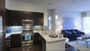 A modern kitchen with dark cabinets, a microwave, and a stove. There is a dining area with a blue sofa and a coffee table. - Hotel  Victoria (Blantyre)
