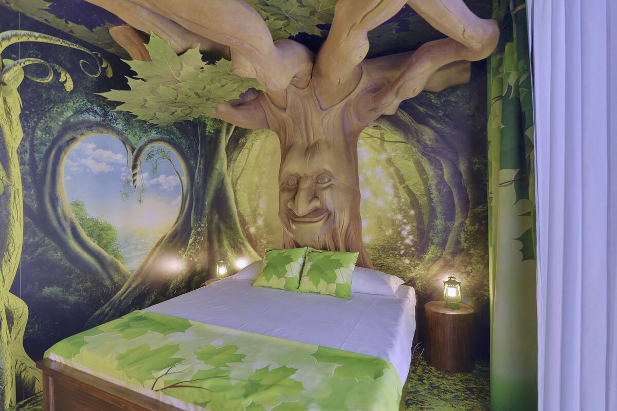 A bedroom with a bed, green leaf-patterned bedding, a tree mural, and two bedside lanterns.
