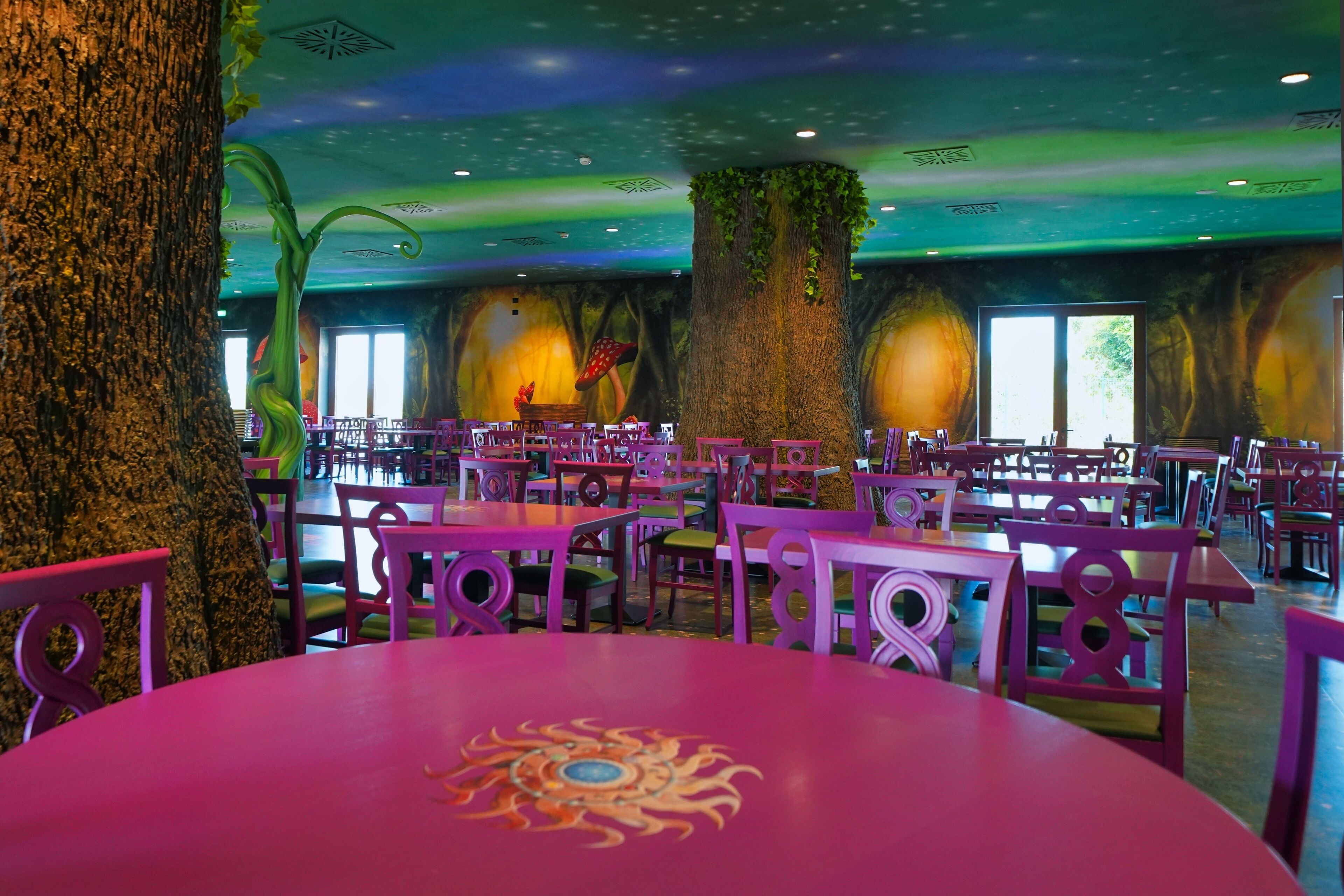 A restaurant with round tables and chairs, large trees, and a mural of a forest on the walls.