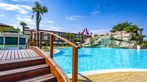 A wooden deck leading to a swimming pool with a clear blue water, surrounded by a paved area and a building with a purple roof in the background.