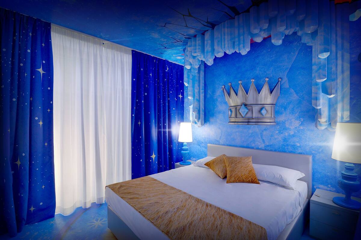A bedroom with a bed, nightstands, a lamp, and a wall with a crown design.