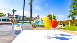 A table with two drinks, a pool, palm trees, and a building in the background.