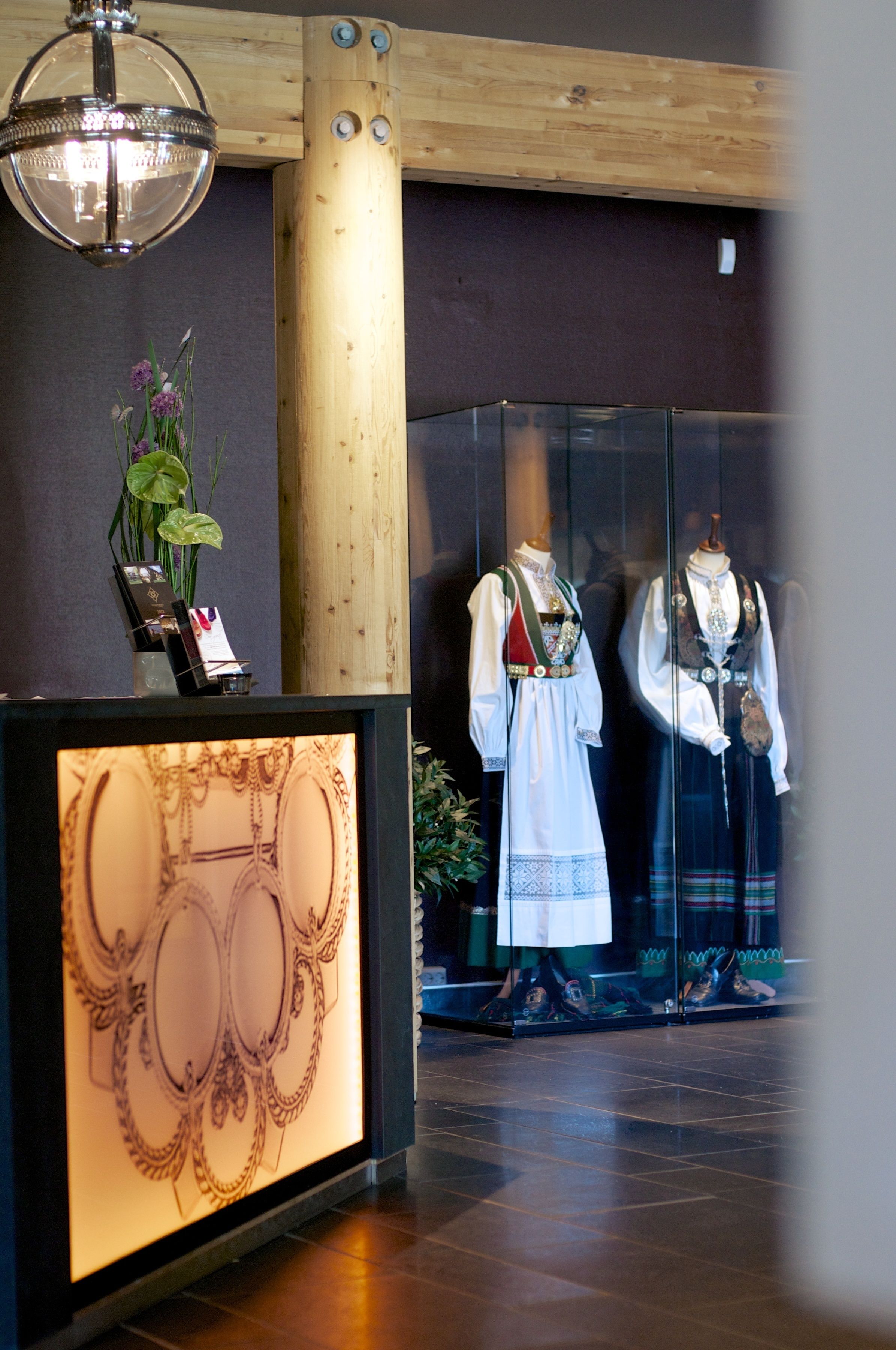 Ett receptionsområde med en träpelare, ett glasmontrinskåp med smycken och skyltdockor klädda i traditionella dräkter.