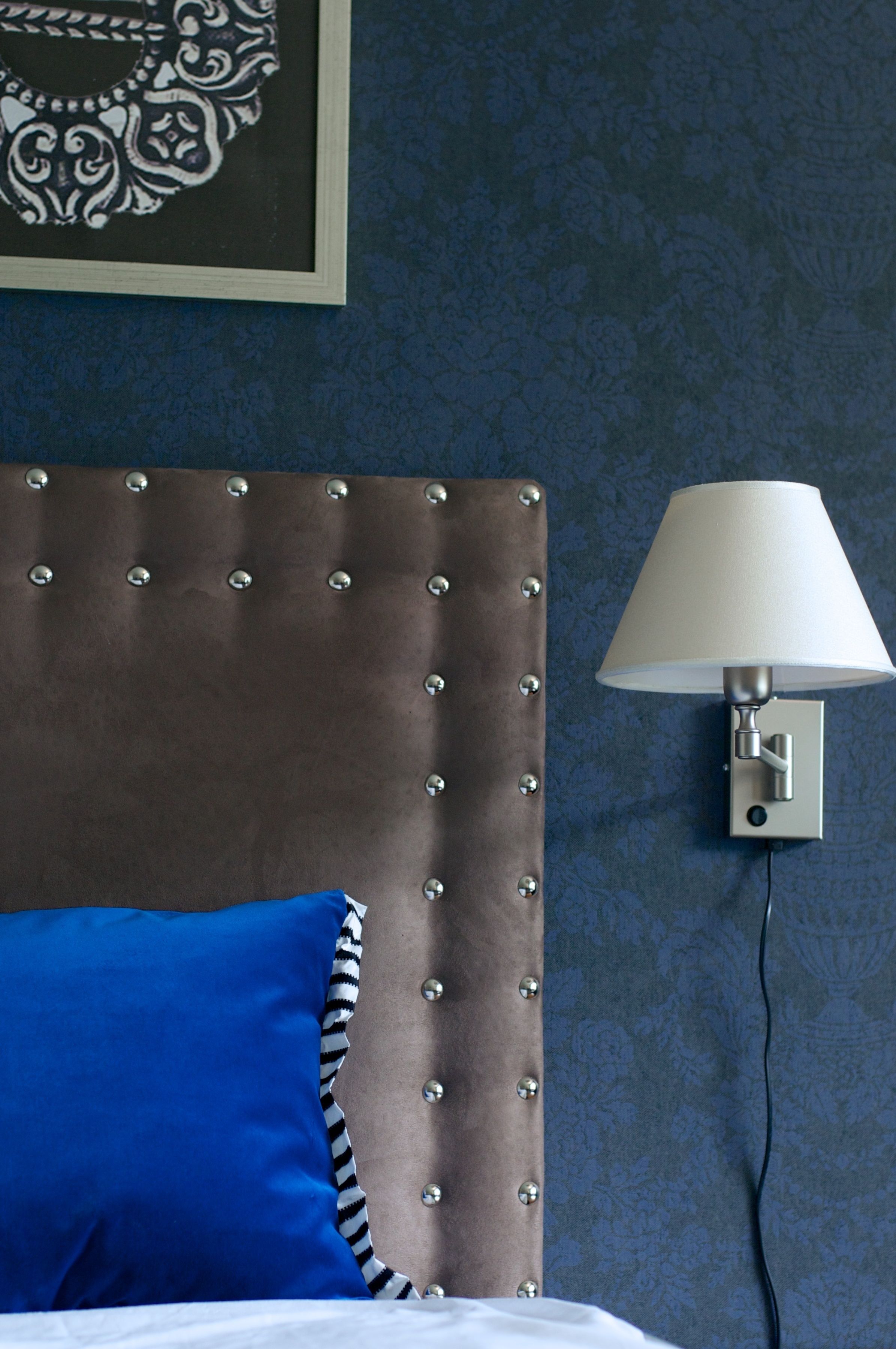 A bedroom with a tufted headboard, a blue pillow, and a wall-mounted lamp.