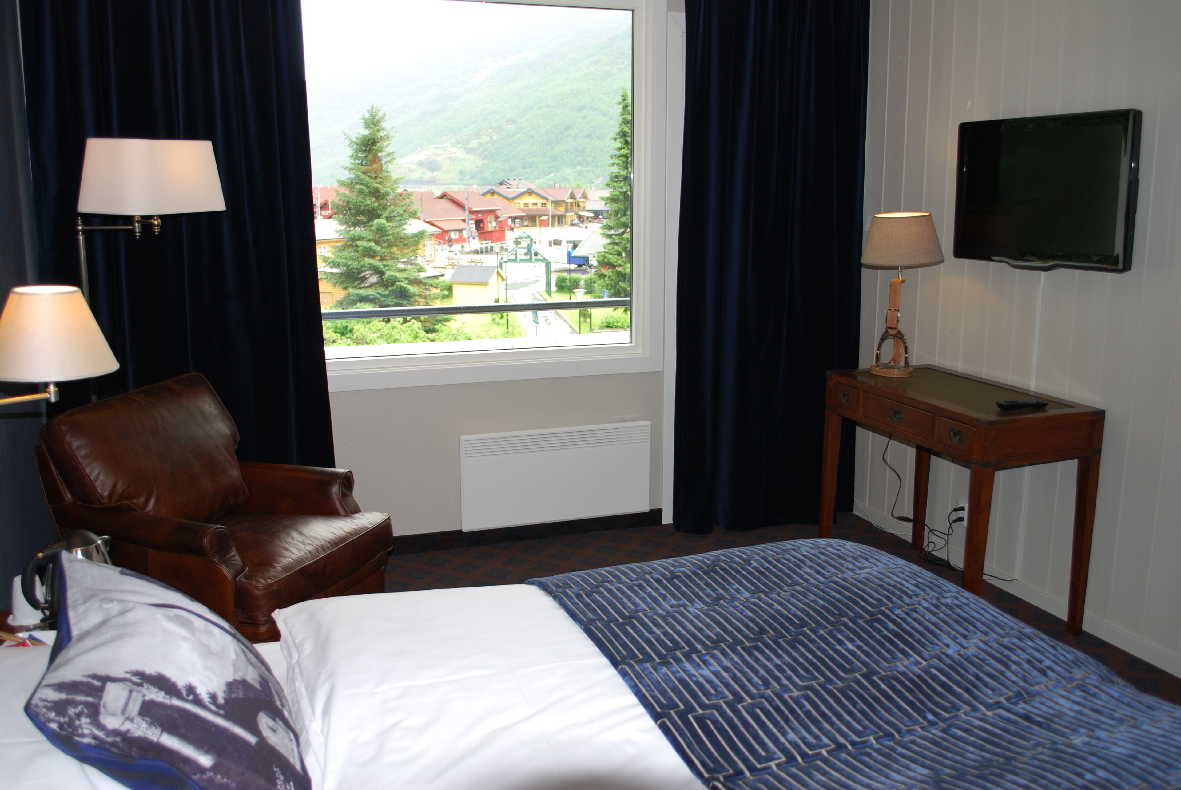A hotel room with a bed, a leather chair, a table with a lamp, a television, and a window with a view of buildings and trees.