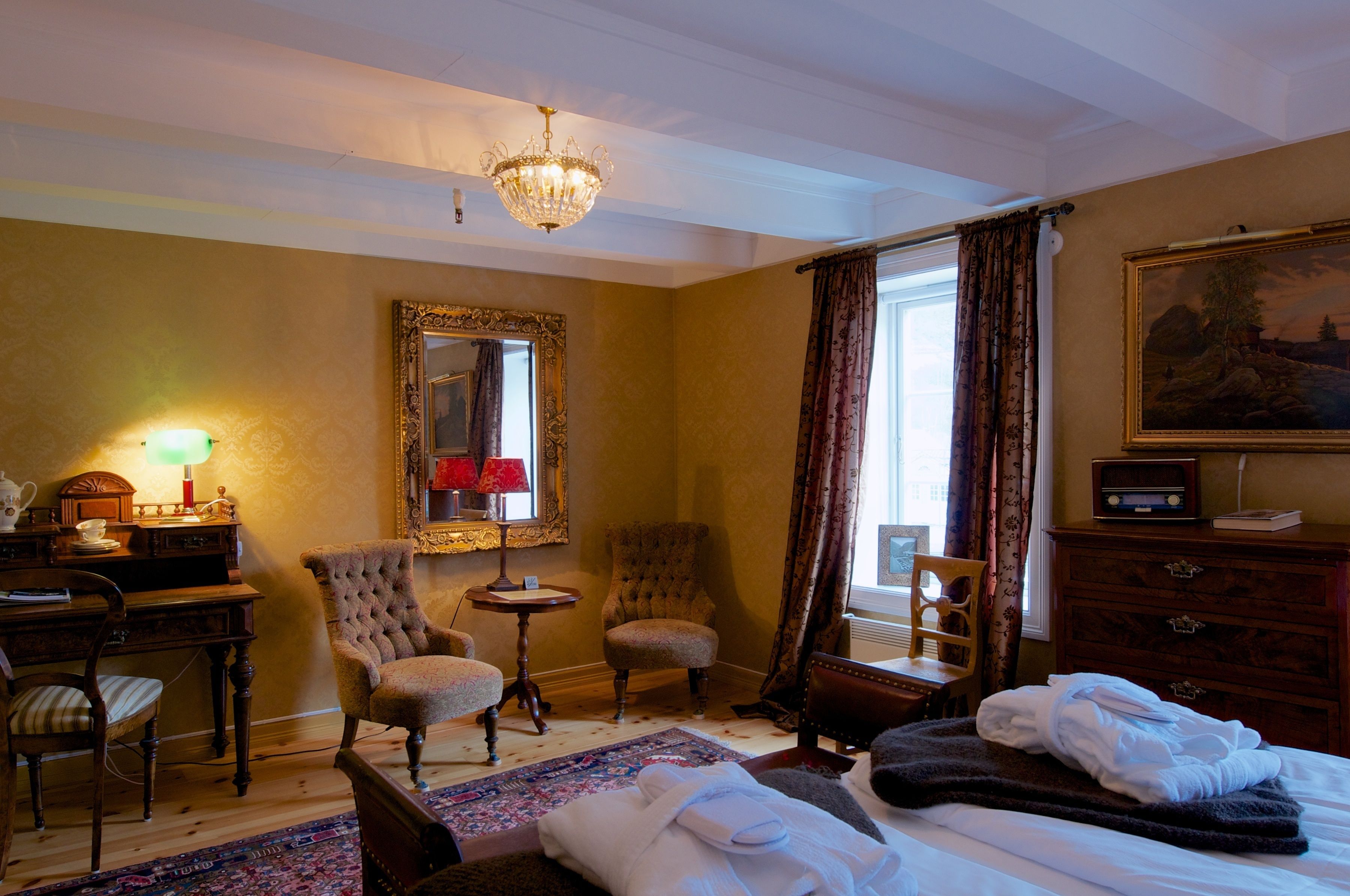 A hotel room with a bed, two armchairs, a small table, a desk with a lamp, a dresser, and a large mirror.